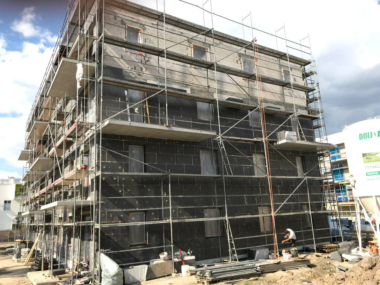 Budynek w trakcie ocieplania grafitowym styropianem, widoczne rusztowania i przygotowania do warstwy zbrojonej. Na dole stos materiałów budowlanych i pracownik.