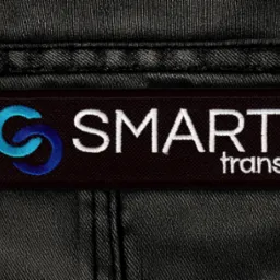 Czarna naszywka z logo 'SMART trans' na tle ciemnego materiału. Logo w kolorach błękitu i bieli, starannie wyhaftowane. Detal odzieży roboczej.