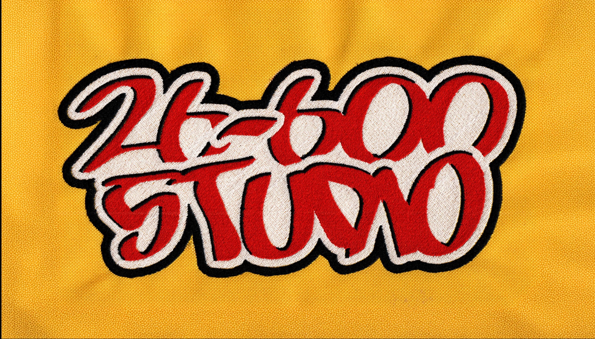 Żółty materiał z wyhaftowanym czerwono-białym napisem 'Action Studio' z czarnym obrysem. Detal haftu na tkaninie.