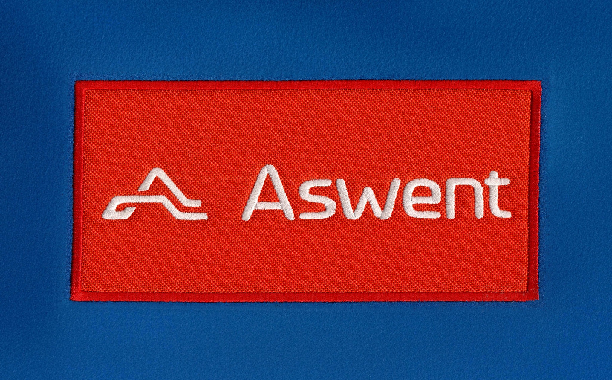 Czerwona naszywka z białym haftowanym logo 'Aswent' na niebieskim tle materiału. Detal odzieży z bliska, precyzyjne wykonanie haftu.