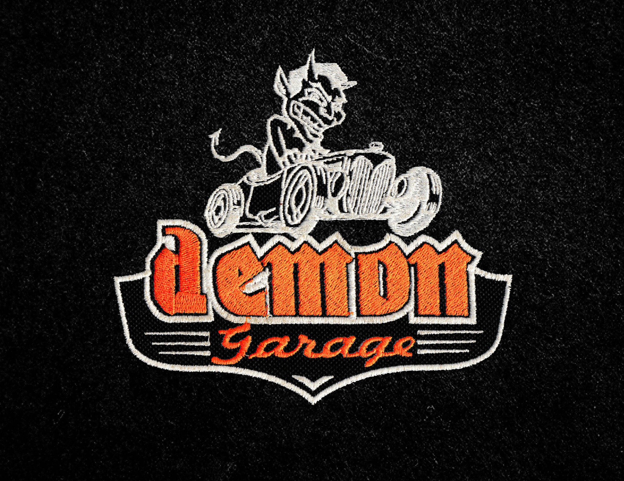 Czarna tkanina z haftowanym, pomarańczowo-białym logo 'Demon Garage' z diabłem w samochodzie. Detal haftu na odzieży.
