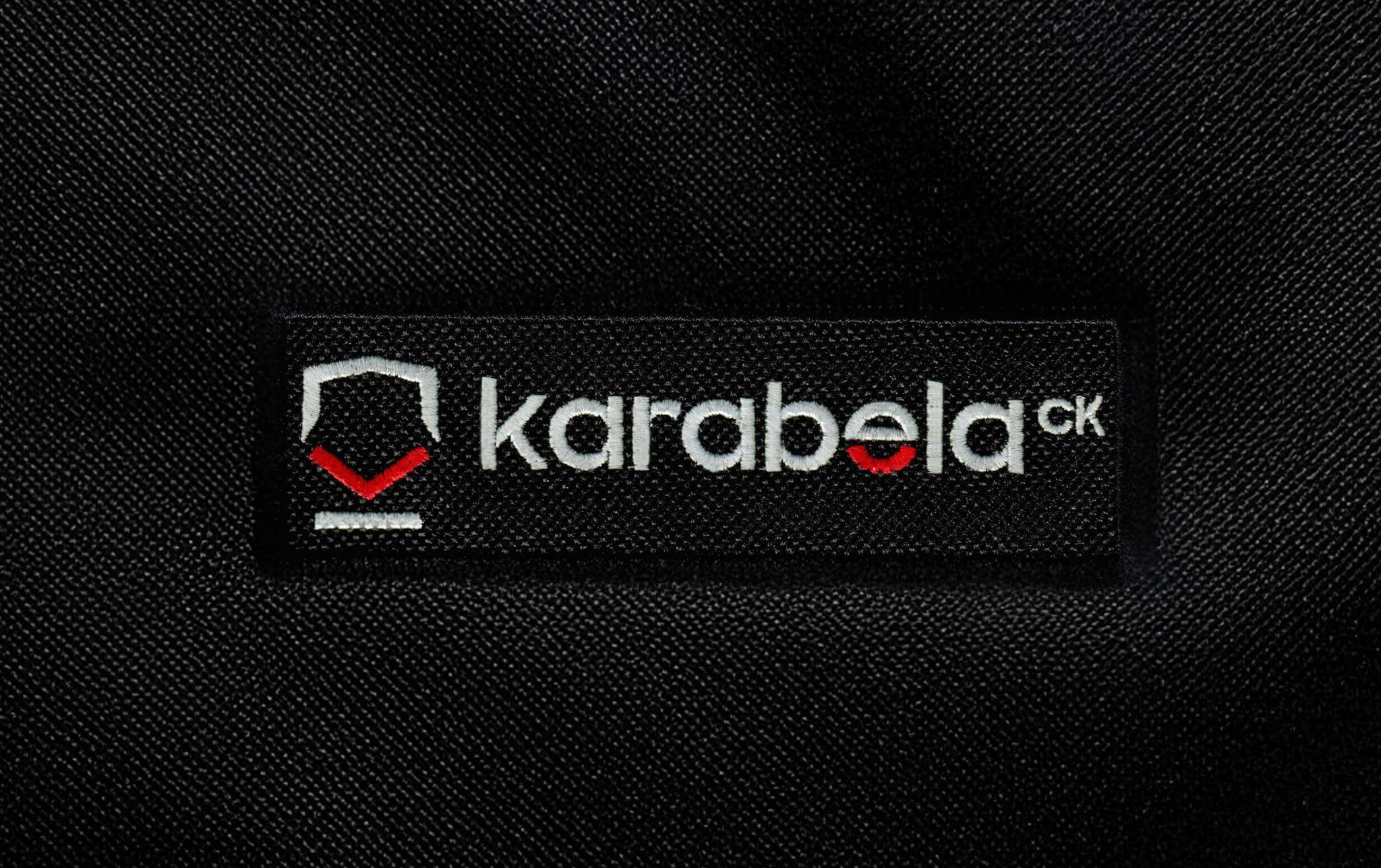 Czarna metka z wyhaftowanym białym logo 'karabelaCK' na tle materiału o wyraźnej fakturze. Czerwony akcent w logo.
