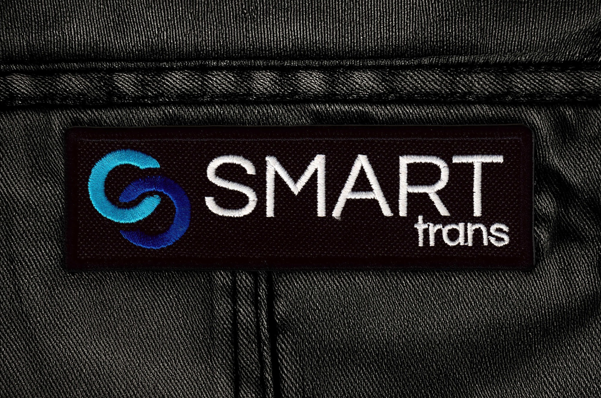 Czarna naszywka z logo 'SMART trans' na tle ciemnego materiału. Logo w kolorach błękitu i bieli, starannie wyhaftowane. Detal odzieży roboczej.