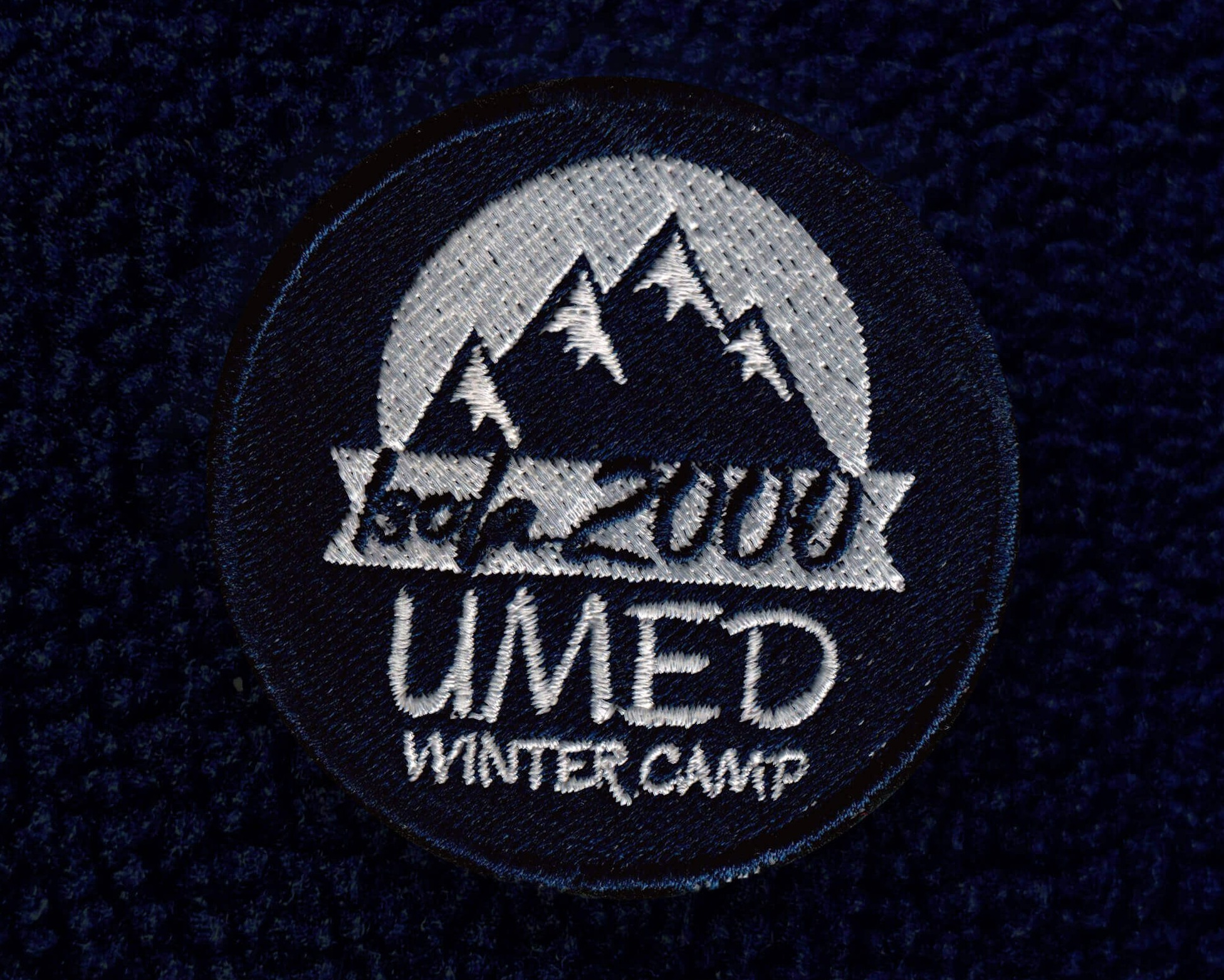 Granatowa naszywka z haftowanym logo 'UMED Winter Camp' z motywem górskim na tle teksturowanej tkaniny, prezentująca detale wykonania.
