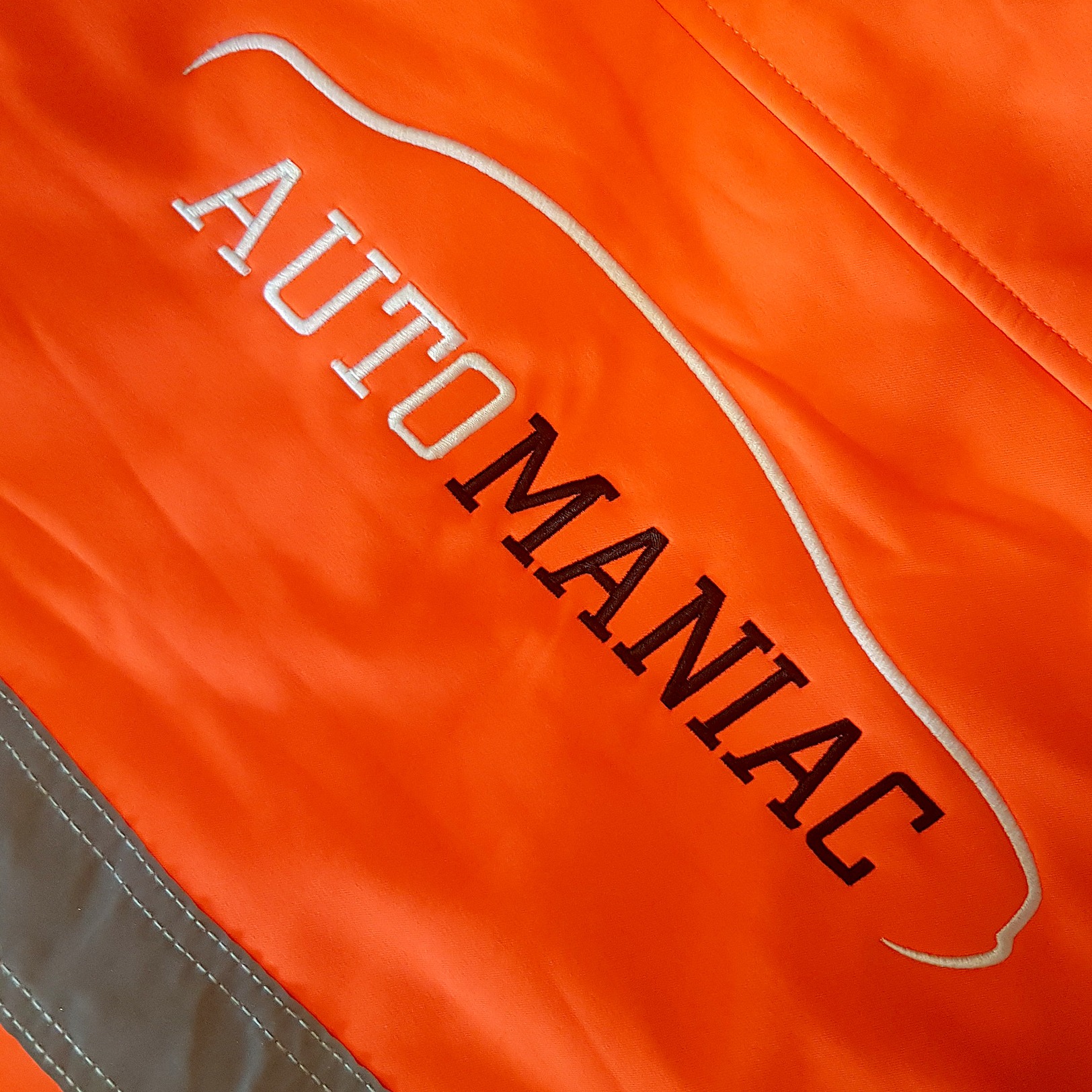 Pomarańczowa odzież robocza z haftowanym logo 'AUTOMANIAC' i białym obrysem grafiki, widoczny fragment szarego pasa odblaskowego.