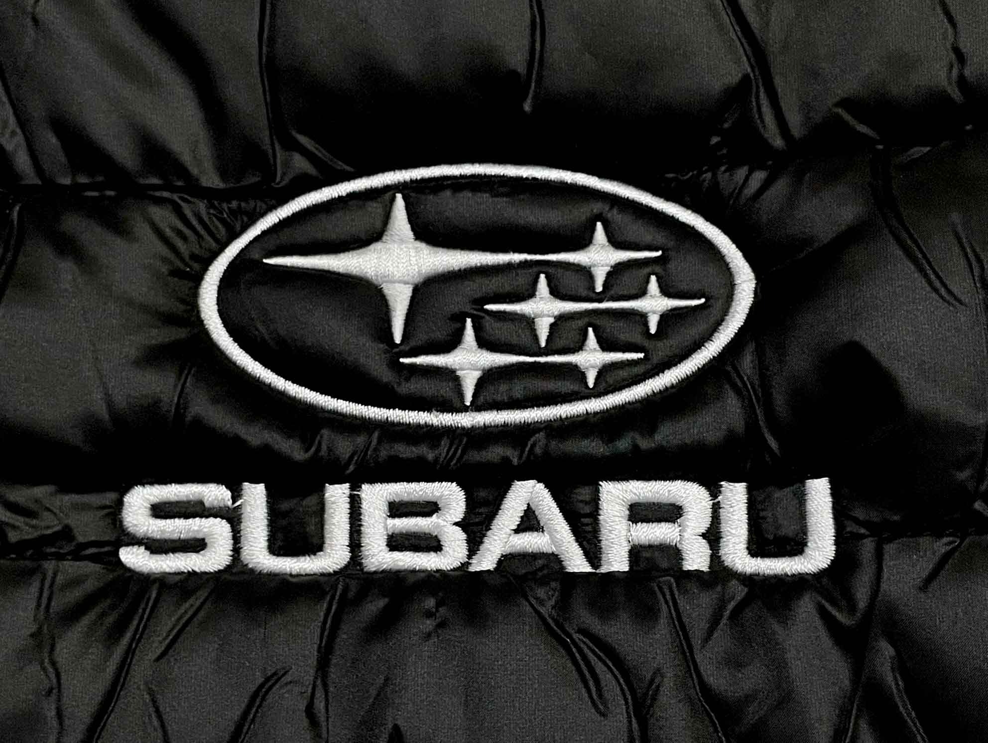 Czarna kurtka puchowa z wyhaftowanym białym logo Subaru na piersi, widoczne detale szwów i tekstury materiału.