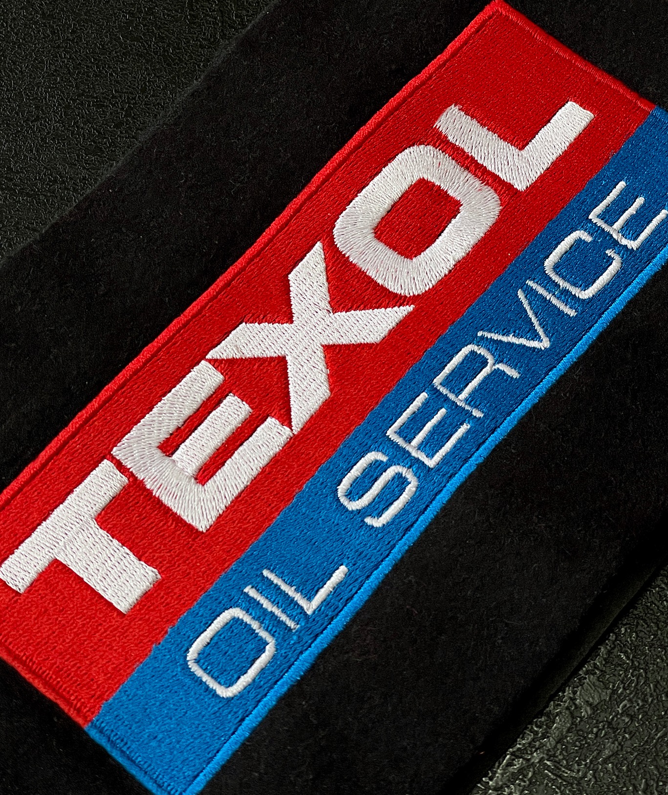 Naszywka z haftowanym logo 'TEXOL OIL SERVICE' na czerwono-niebieskim tle, przyszyta do czarnego materiału. Detal haftu, wyraźne szwy.