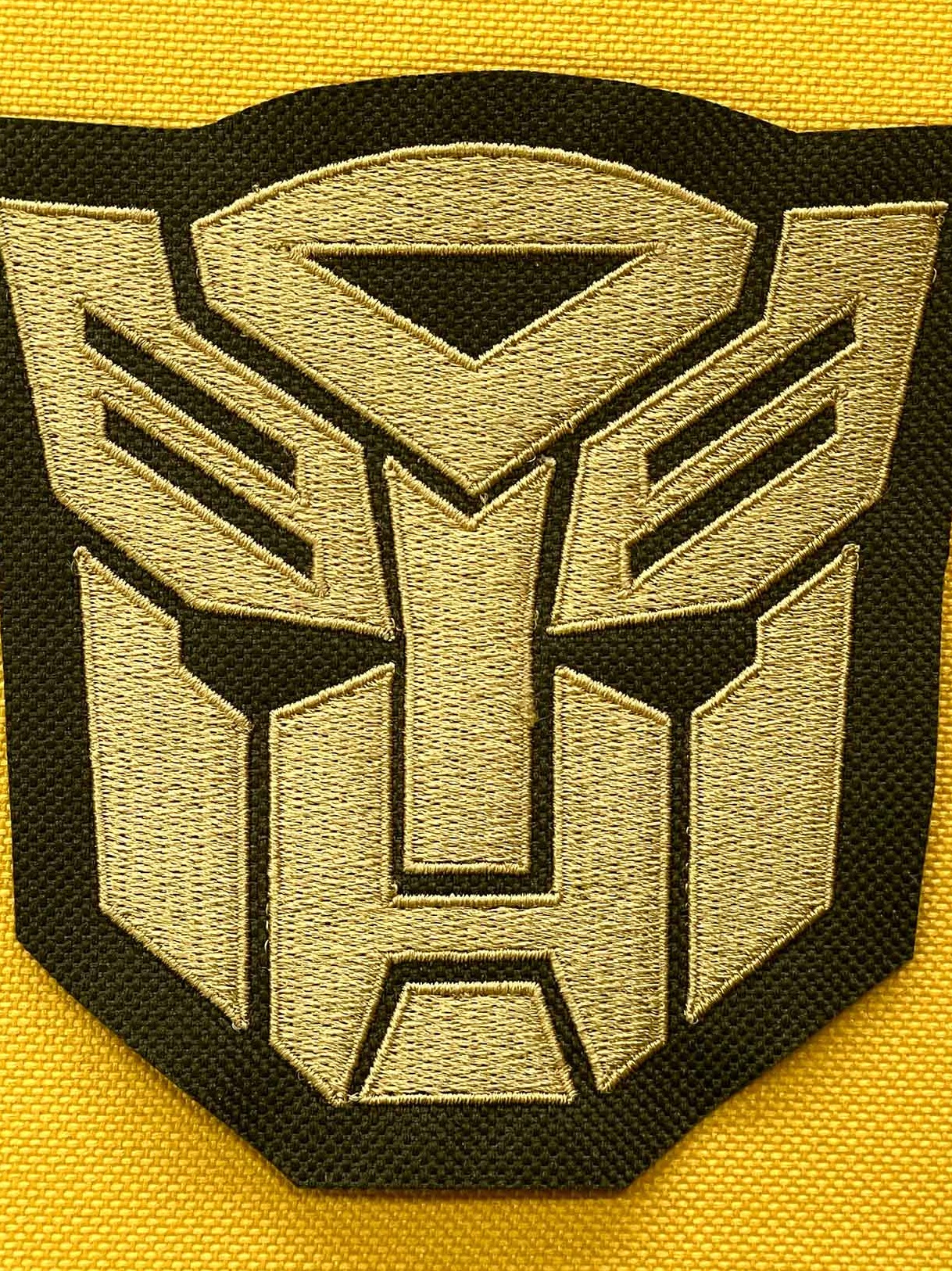 Czarna naszywka z haftowanym złotym logo Transformers na żółtym materiale. Detal haftu, widoczna tekstura tkaniny.