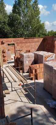 Wznoszenie ścian z czerwonej cegły ceramicznej na budowie domu jednorodzinnego. Widoczne rusztowania i palety z cegłą. Praca w toku, w słoneczny dzień.