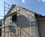 Ocieplony, szary dom jednorodzinny w trakcie prac wykończeniowych. Widoczne rusztowania, drabina, okno bez ramy. Słoneczny dzień, błękitne niebo.