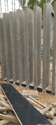 Szereg betonowych słupów konstrukcyjnych w trakcie budowy, widoczne zbrojenia i elementy szalunkowe u podstawy. Ujęcie z dołu, w tle zieleń.