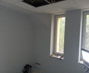 Wnętrze pomalowanego pokoju z dwoma oknami i częściowo podwieszonym sufitem, ukazujące etap wykończenia. Widoczne gniazdka elektryczne i elementy instalacji.