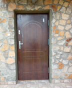 Brązowe drzwi wejściowe z metalowymi okuciami, osadzone w ścianie z nieregularnego kamienia, z widoczną instalacją elektryczną po lewej stronie.