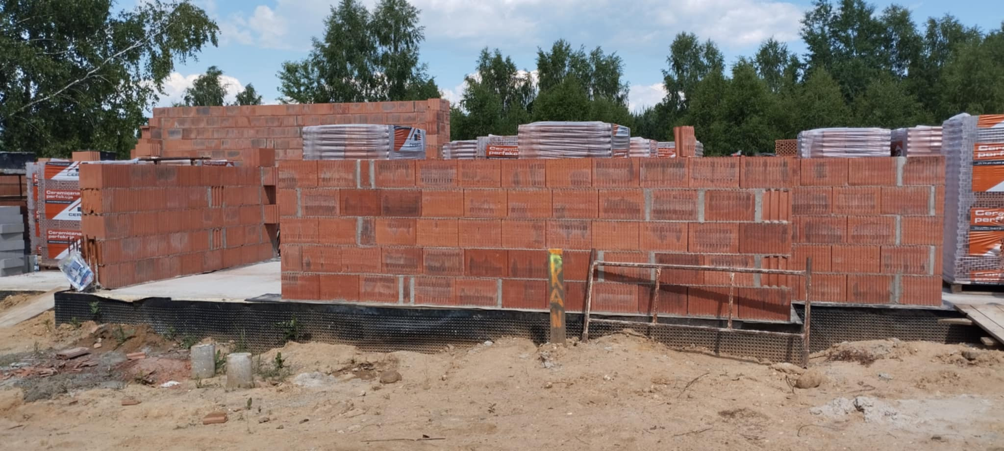 Wznoszenie ścian z czerwonej cegły ceramicznej na fundamencie. Widoczne palety z cegłami i zarys lasu w tle. Etap budowy domu jednorodzinnego.