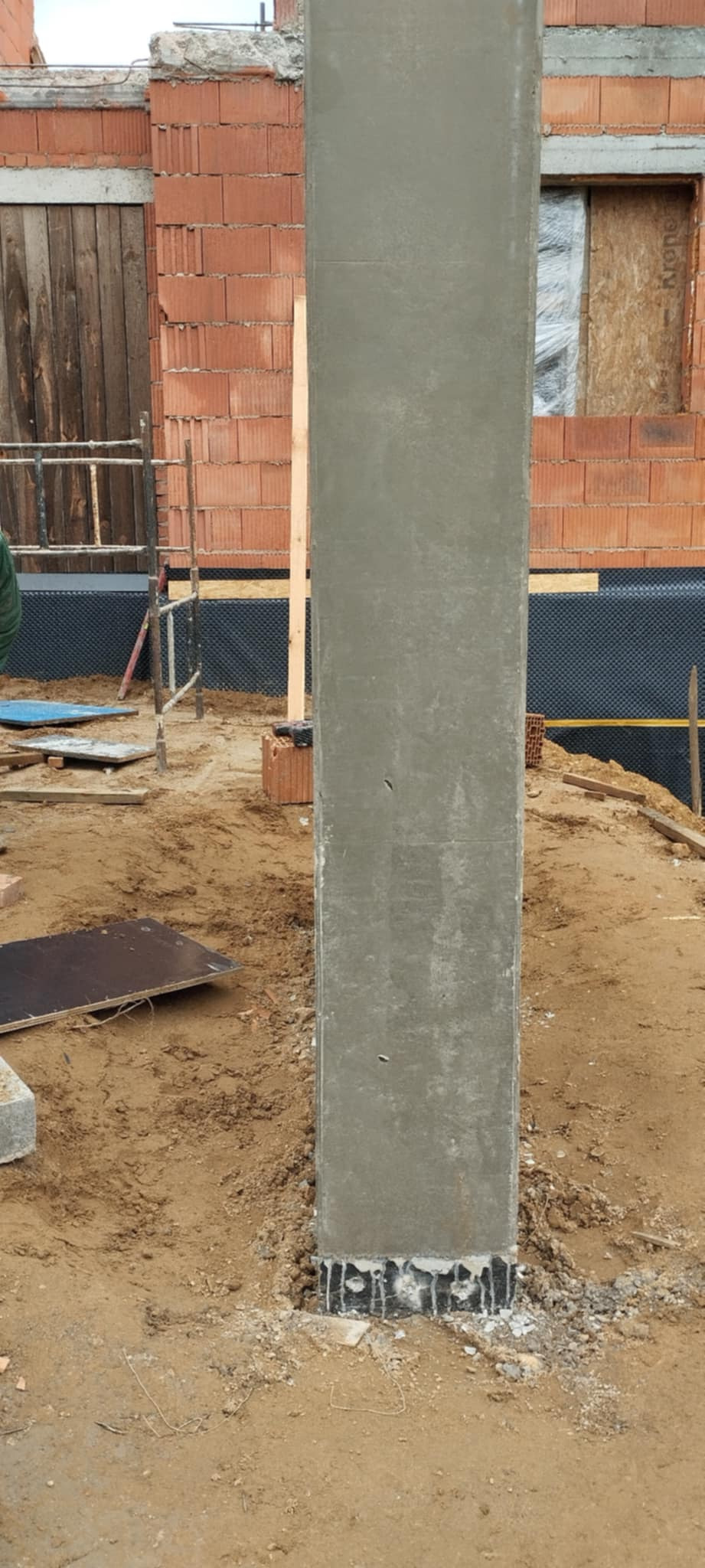 Żelbetowy słup w trakcie budowy domu jednorodzinnego. Widoczne szalunki, cegły i elementy konstrukcyjne. Otoczenie budowy, piasek.