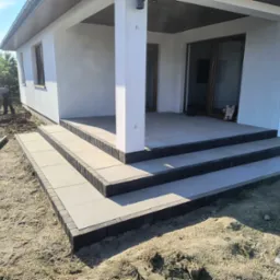 Nowoczesne schody wejściowe z płytek i kostki brukowej przy budynku w Gostyninie. Widoczna elewacja i prace ziemne w tle. Jasne barwy, minimalistyczny design.