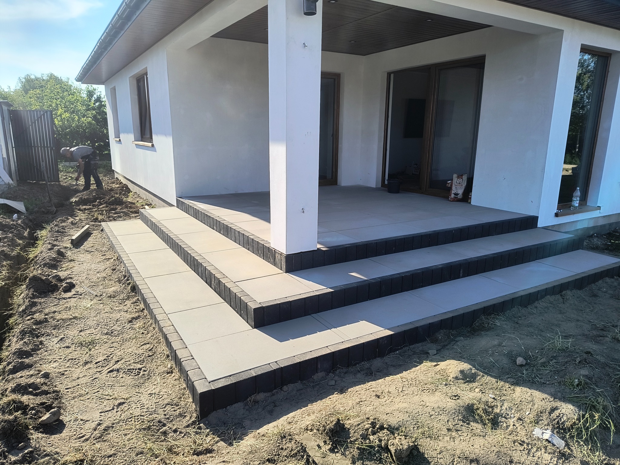 Nowoczesne schody wejściowe z płytek i kostki brukowej przy budynku w Gostyninie. Widoczna elewacja i prace ziemne w tle. Jasne barwy, minimalistyczny design.