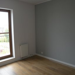 House color renovation - Pomalowane na szaro wnętrze pokoju z drewnianą podłogą, białym grzejnikiem i oknem z widokiem na zieleń. Świeżo wykończone, minimalistyczne wnętrze.