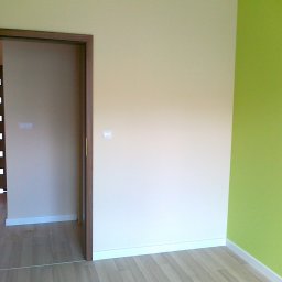 House color renovation - Pokój po odświeżeniu: kremowa ściana z kontaktem, zielona ściana narożna, drewniana podłoga i drzwi z brązową ościeżnicą. Widok na sąsiednie pomieszczenie.