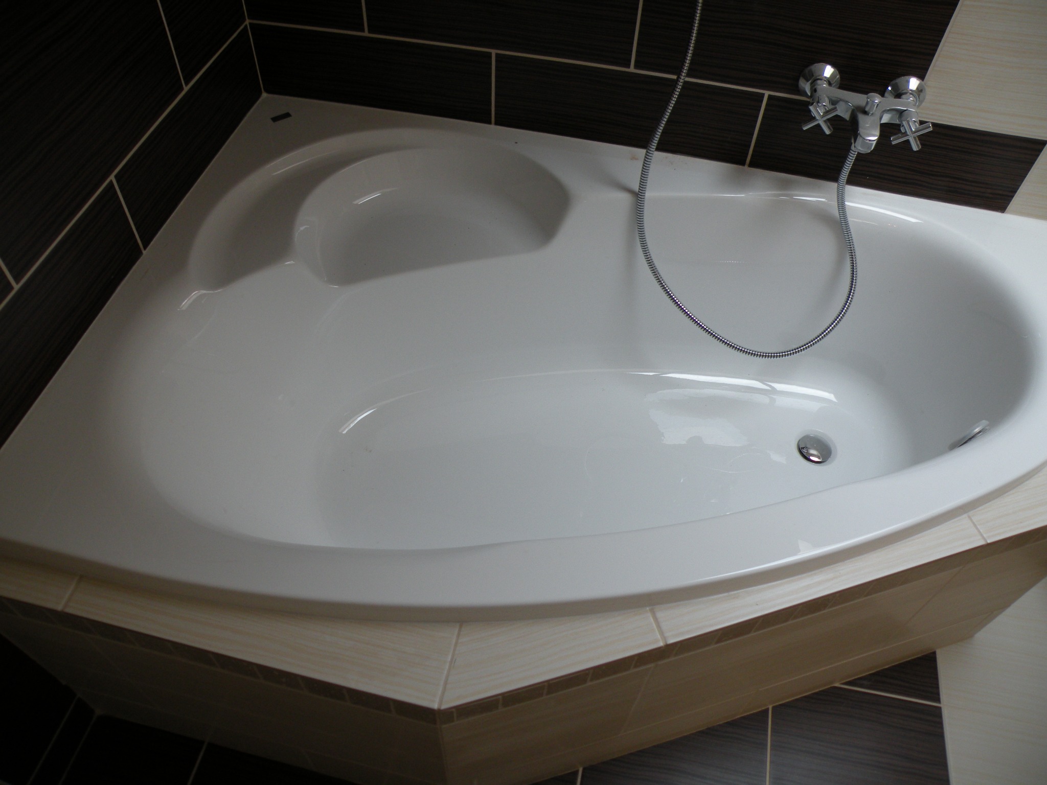Biała, narożna wanna z chromowaną baterią i wężem, obudowana beżowymi płytkami. W tle ciemne, brązowe płytki na ścianie. Widok z góry.