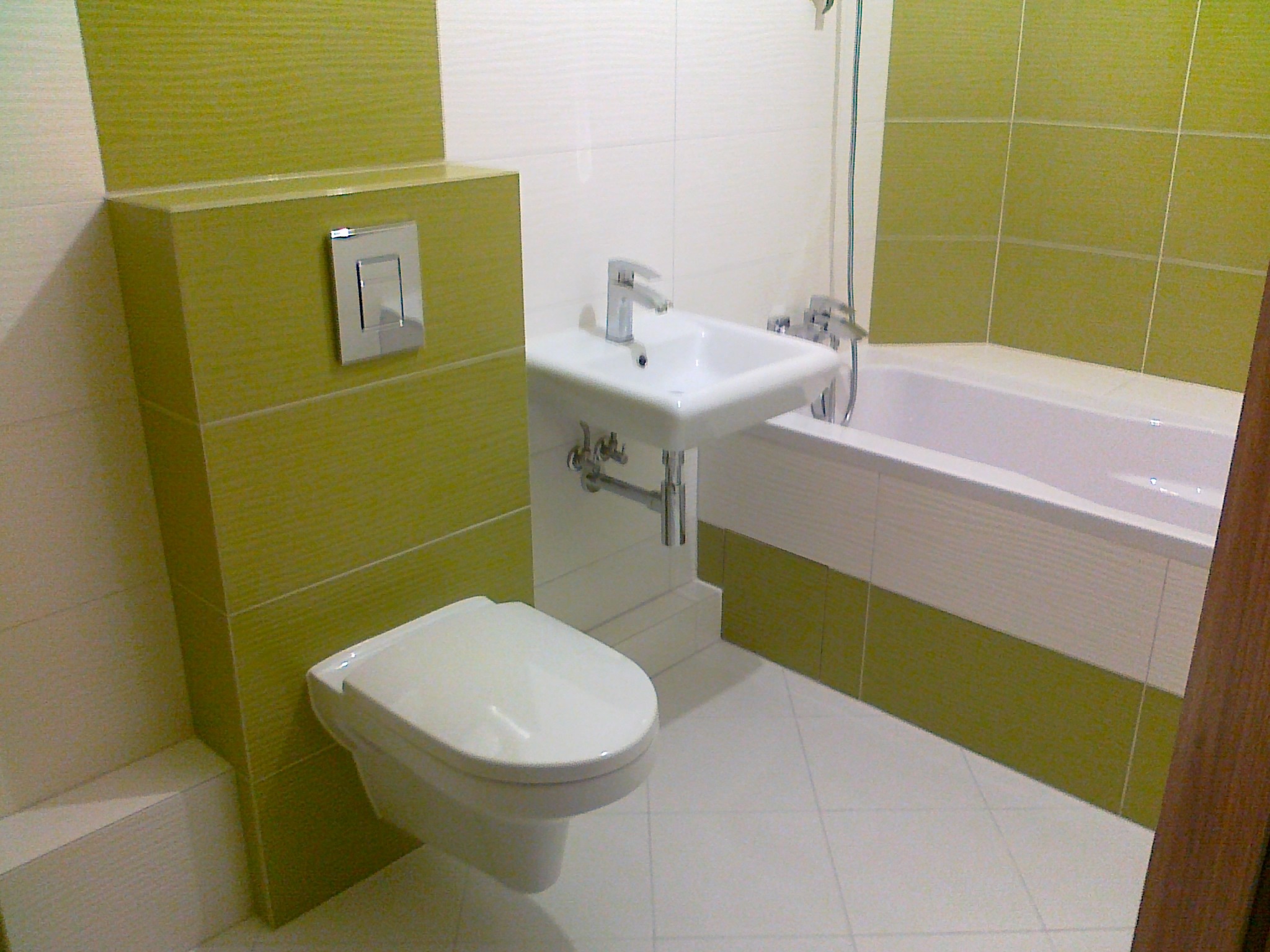 Wykończona łazienka z białą armaturą i zielonymi akcentami. Umywalka na postumencie, podwieszana toaleta i wanna z prysznicem. Białe płytki podłogowe i częściowo na ścianach.