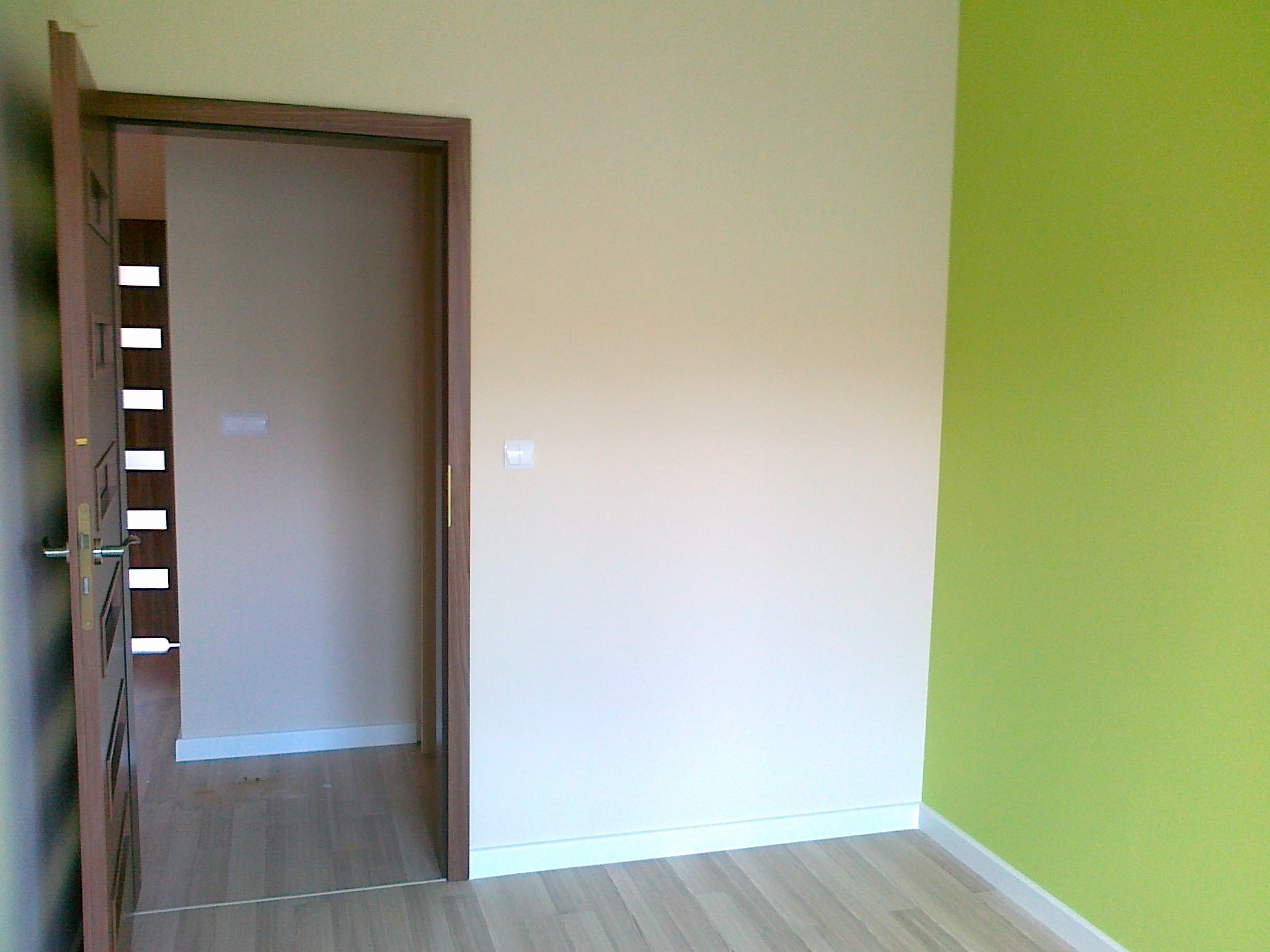 Pokój po odświeżeniu: kremowa ściana z kontaktem, zielona ściana narożna, drewniana podłoga i drzwi z brązową ościeżnicą. Widok na sąsiednie pomieszczenie.