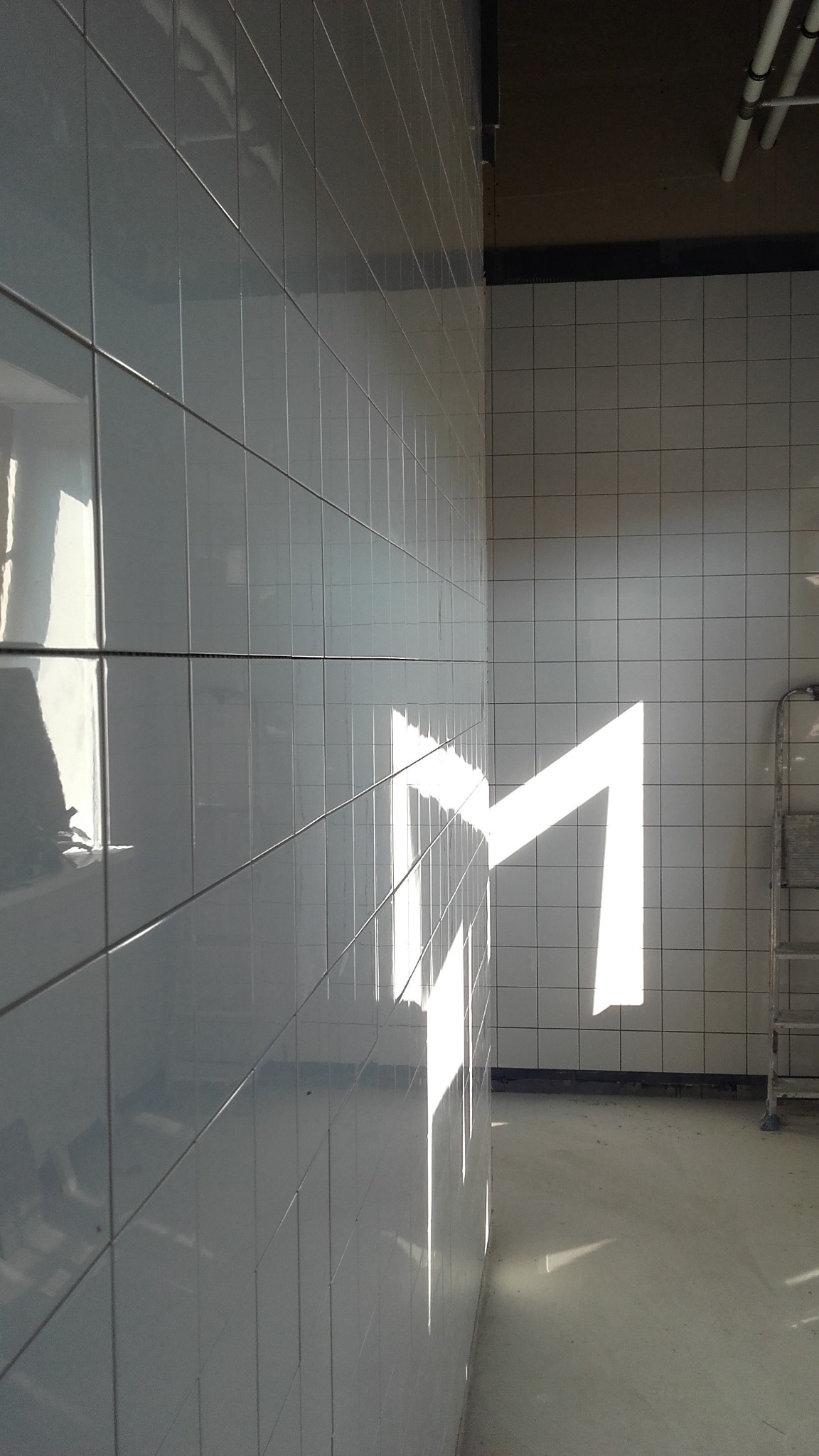 Błyszczące, białe płytki ścienne z widocznym załamaniem światła, tworzącym geometryczny wzór. W tle fragment ściany z mniejszymi płytkami i drabina.