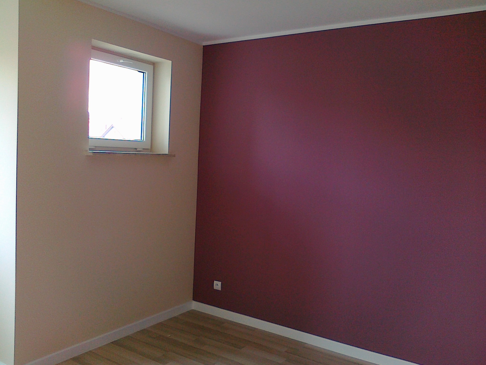 Pokój po malowaniu: ściana w odcieniu burgundu, druga w beżu, okno z białymi ramami, jasna podłoga. Widoczny kontakt elektryczny.