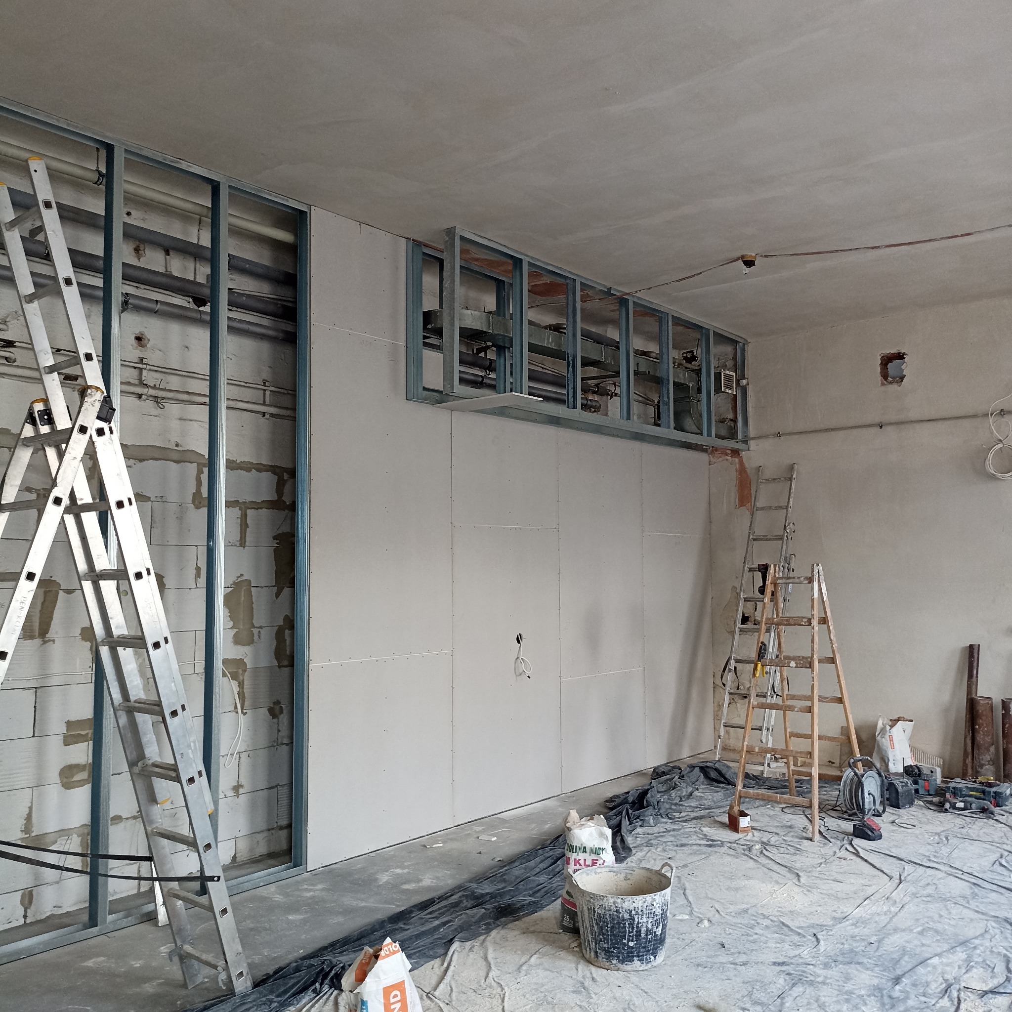 Montaż płyt gipsowo-kartonowych na metalowej konstrukcji w pomieszczeniu. Widoczne drabiny, narzędzia i materiały budowlane. Prace wykończeniowe w surowym wnętrzu.