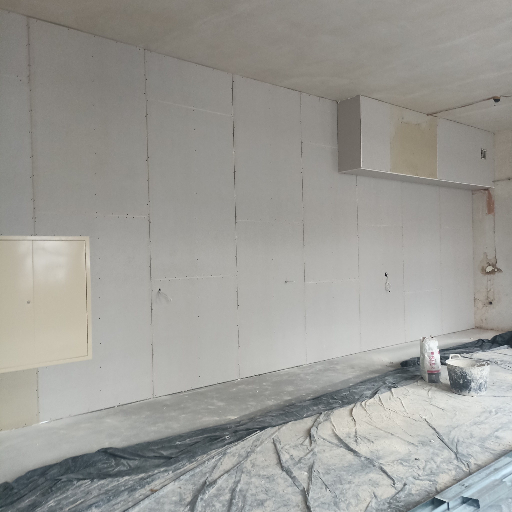 Ściana z płyt kartonowo-gipsowych w trakcie montażu, widoczne łączenia płyt i narzędzia pracy. Częściowo zakryta podłoga folią ochronną. W tle surowy stan wykończenia.