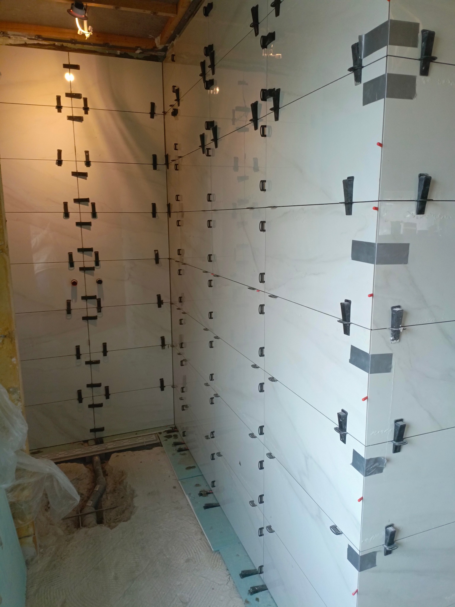 Białe płytki imitujące marmur, ułożone na ścianie z użyciem klipsów poziomujących i sznurka, widok na narożnik łazienki w trakcie prac wykończeniowych.