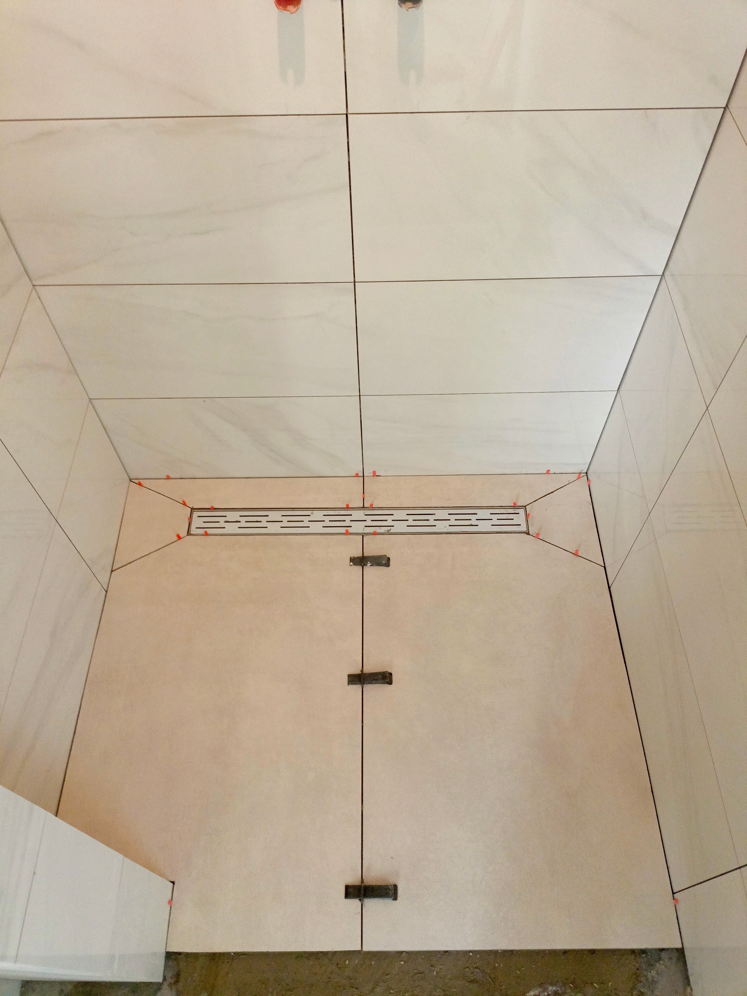 Jasne, kwadratowe płytki ułożone na podłodze i ścianach. Widoczny odpływ liniowy i krzyżyki dystansowe. Ujęcie z góry ukazuje postęp prac w łazience.