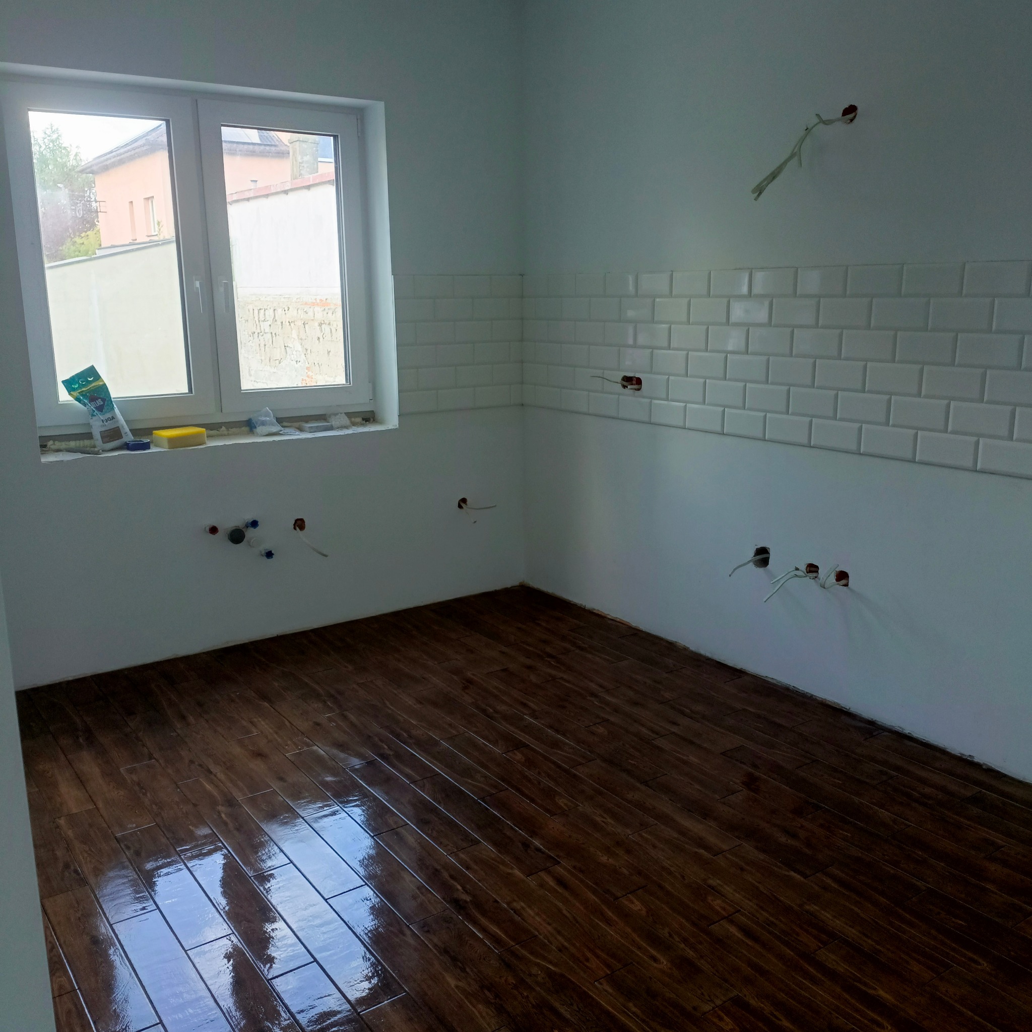 Wnętrze w trakcie remontu: ciemna, drewnopodobna podłoga, białe ściany z ułożonymi białymi płytkami typu metro, okno z widokiem na zewnątrz i wystające kable.