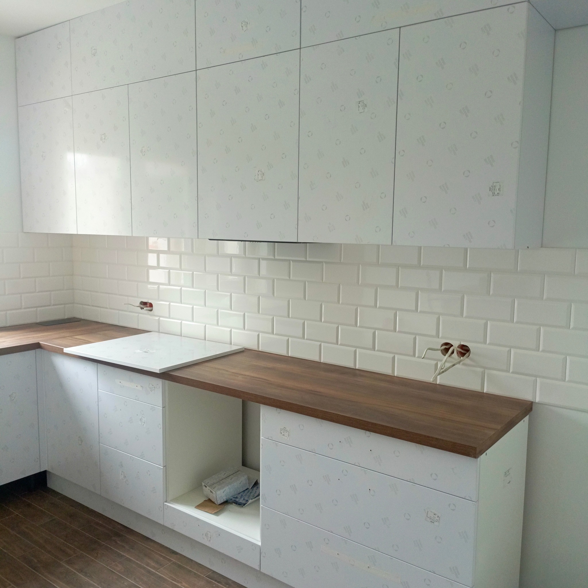 Kuchnia w trakcie wykańczania: białe szafki z folią ochronną, drewniany blat, białe płytki metro na ścianie. Widoczne niedokończone gniazdka.