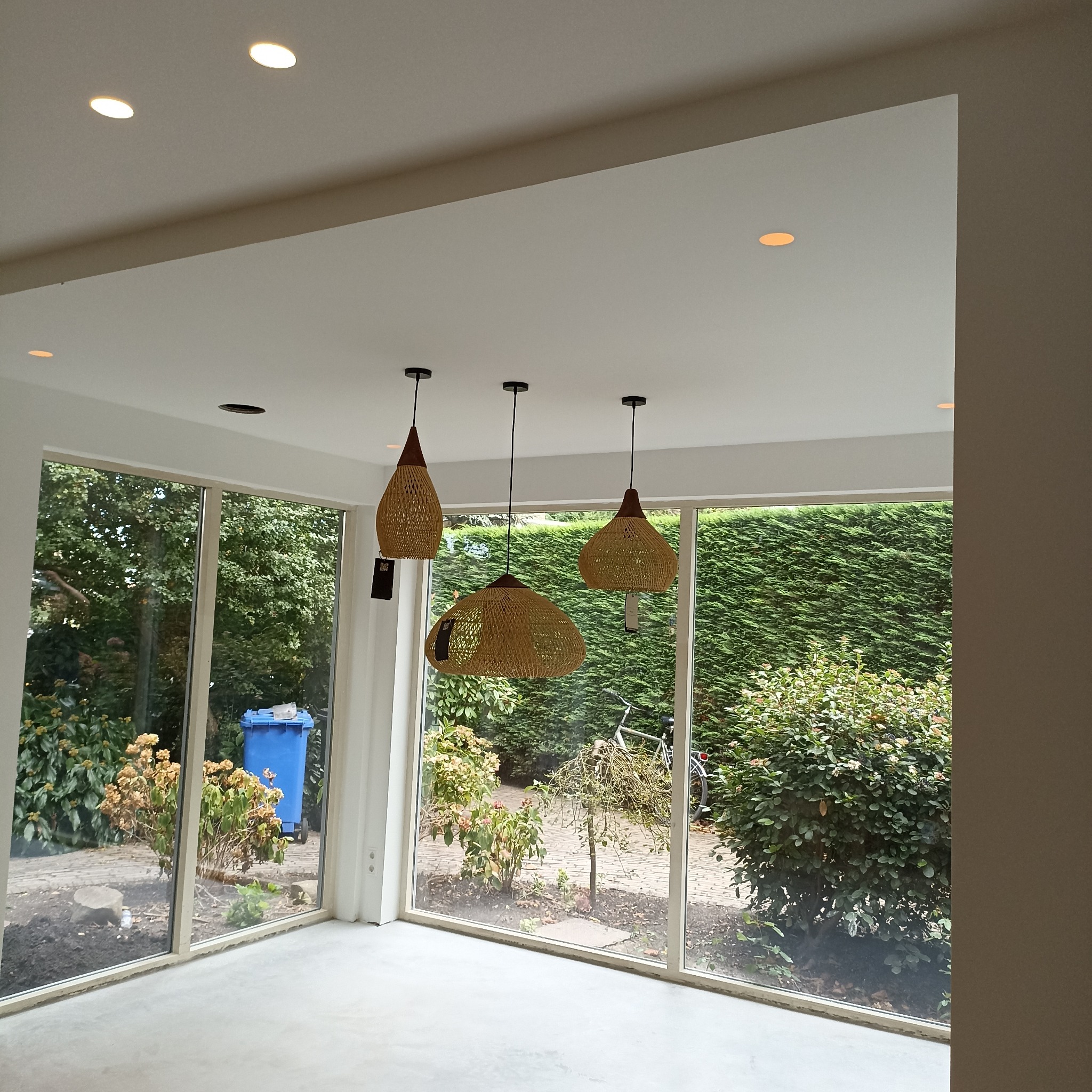 Jasne wnętrze z trzema plecionymi lampami wiszącymi, widok na ogród przez duże okna. Nowoczesne oświetlenie punktowe w suficie.