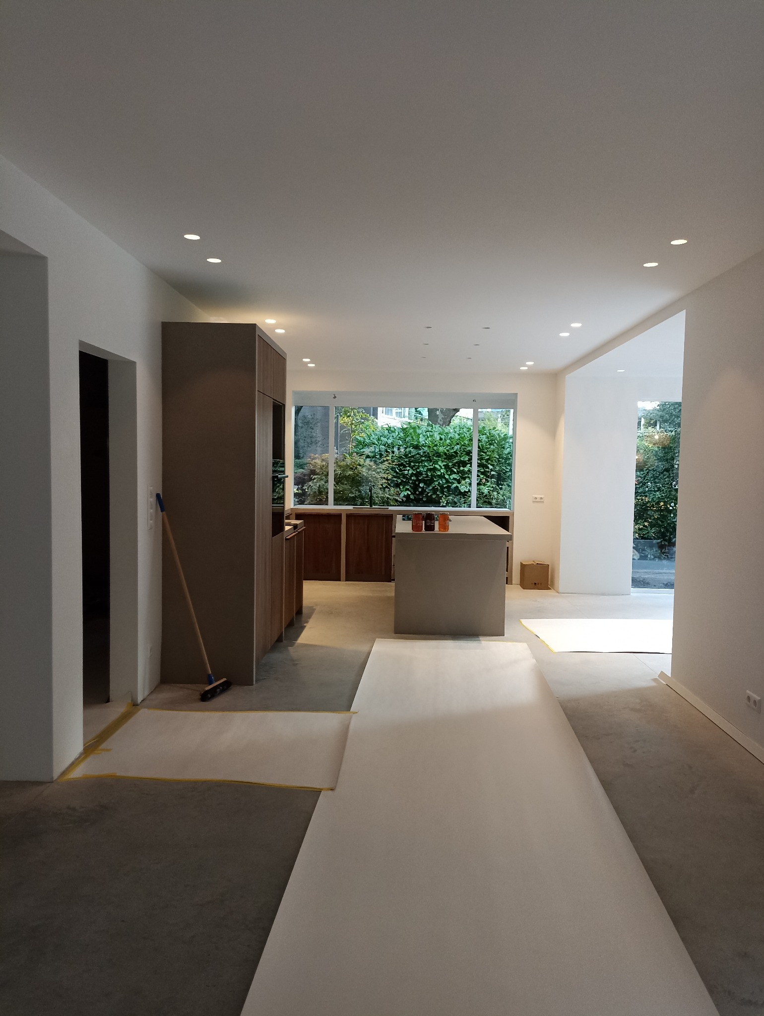 Wnętrze w trakcie prac wykończeniowych: zabezpieczona podłoga, zabudowa kuchenna w tle, widoczne okno z zielenią. Świeże, jasne ściany.