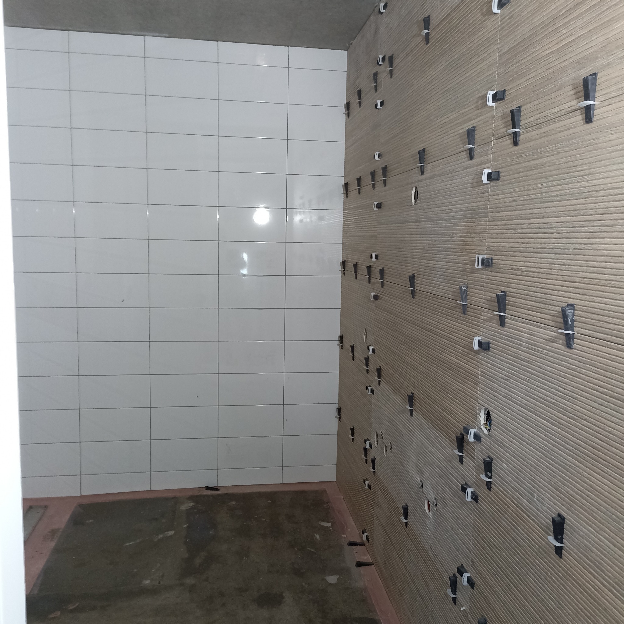 Łazienka w trakcie prac: białe płytki na ścianie obok brązowych, ryflowanych płytek z systemem poziomowania. Widoczny surowy betonowy sufit i podłoga w remoncie.