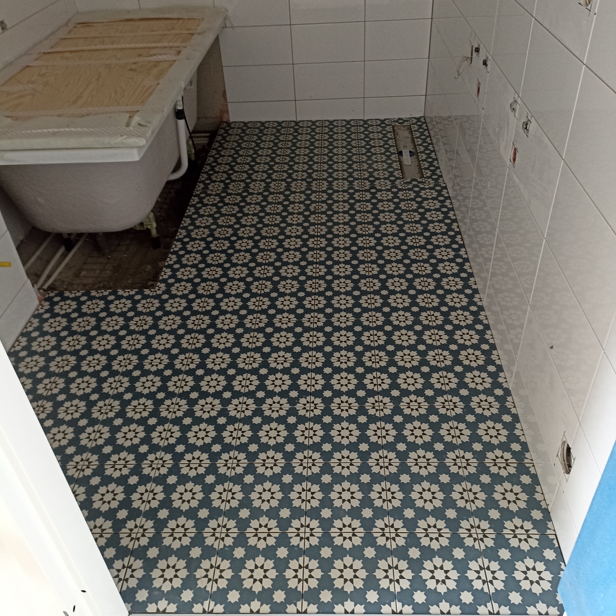 Wykończona podłoga w łazience z dekoracyjnymi, niebiesko-białymi płytkami. Widoczna wanna z zabezpieczeniem i białe płytki ścienne. Gniazdka elektryczne bez osprzętu.