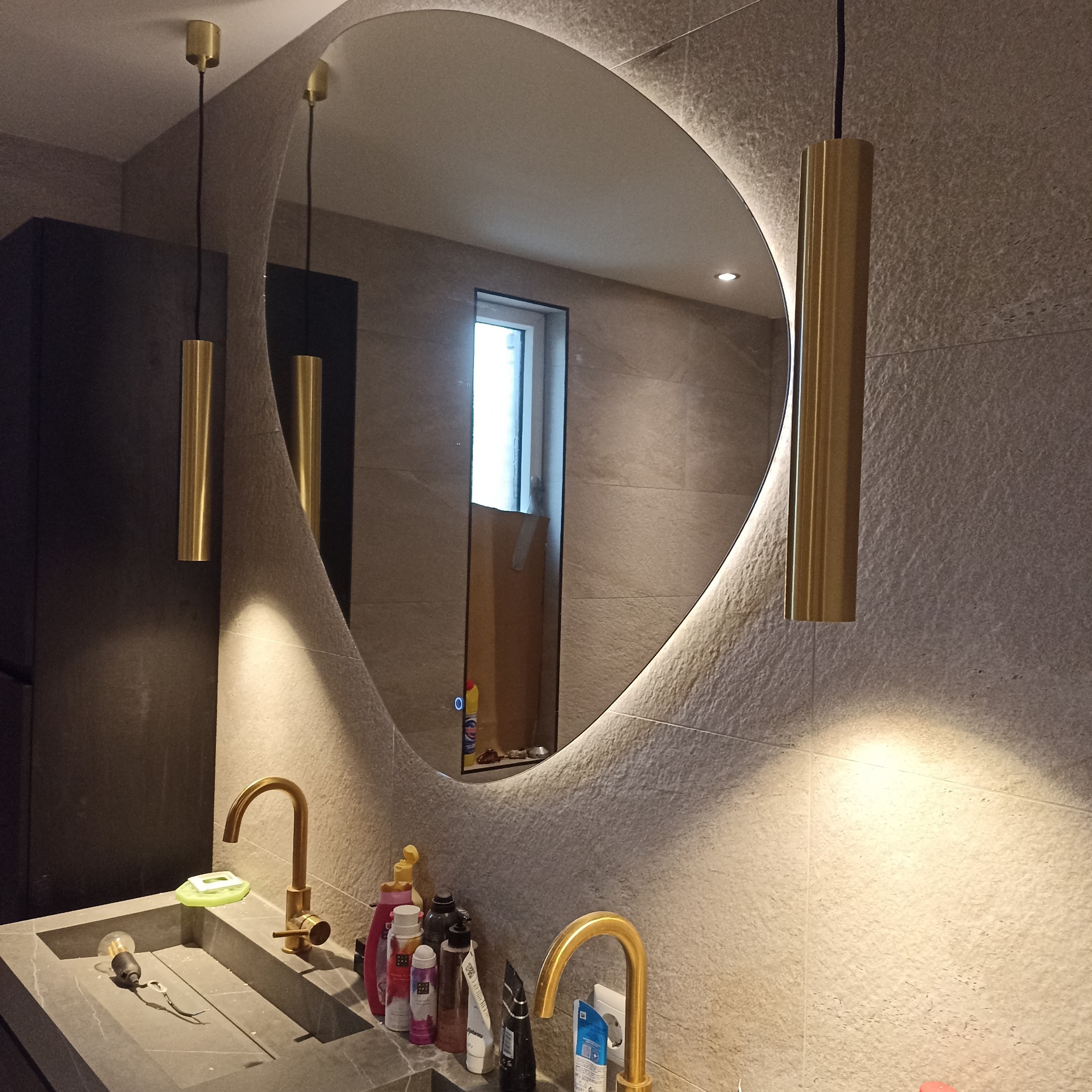 Nowoczesna łazienka z owalnym lustrem z podświetleniem LED, złotymi lampami wiszącymi i kamienną umywalką. Minimalistyczny design, szare płytki, eleganckie wykończenie.