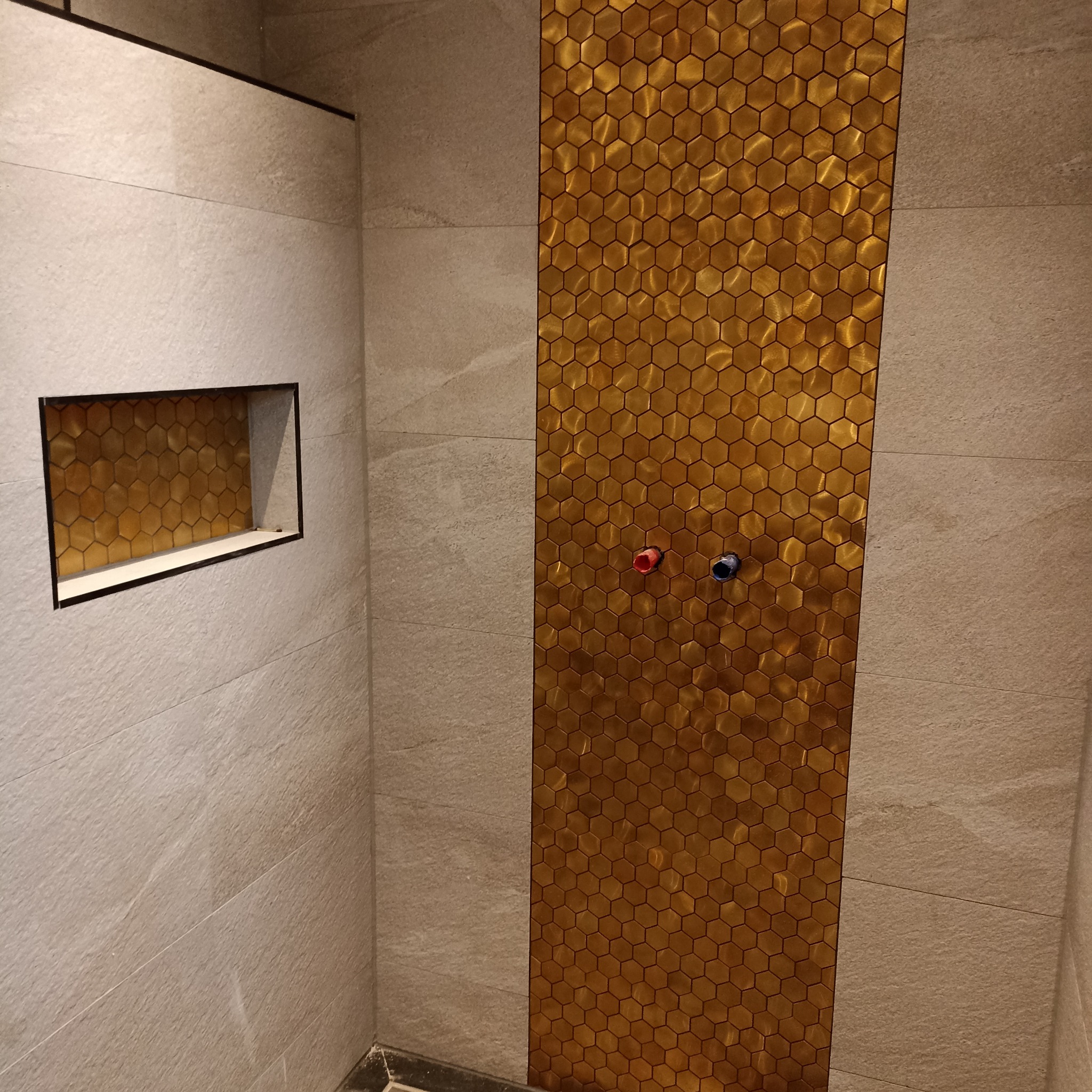 Beżowe płytki w kabinie prysznicowej z wnęką i pionowym pasem złotych heksagonalnych płytek, widoczne przyłącza wody.