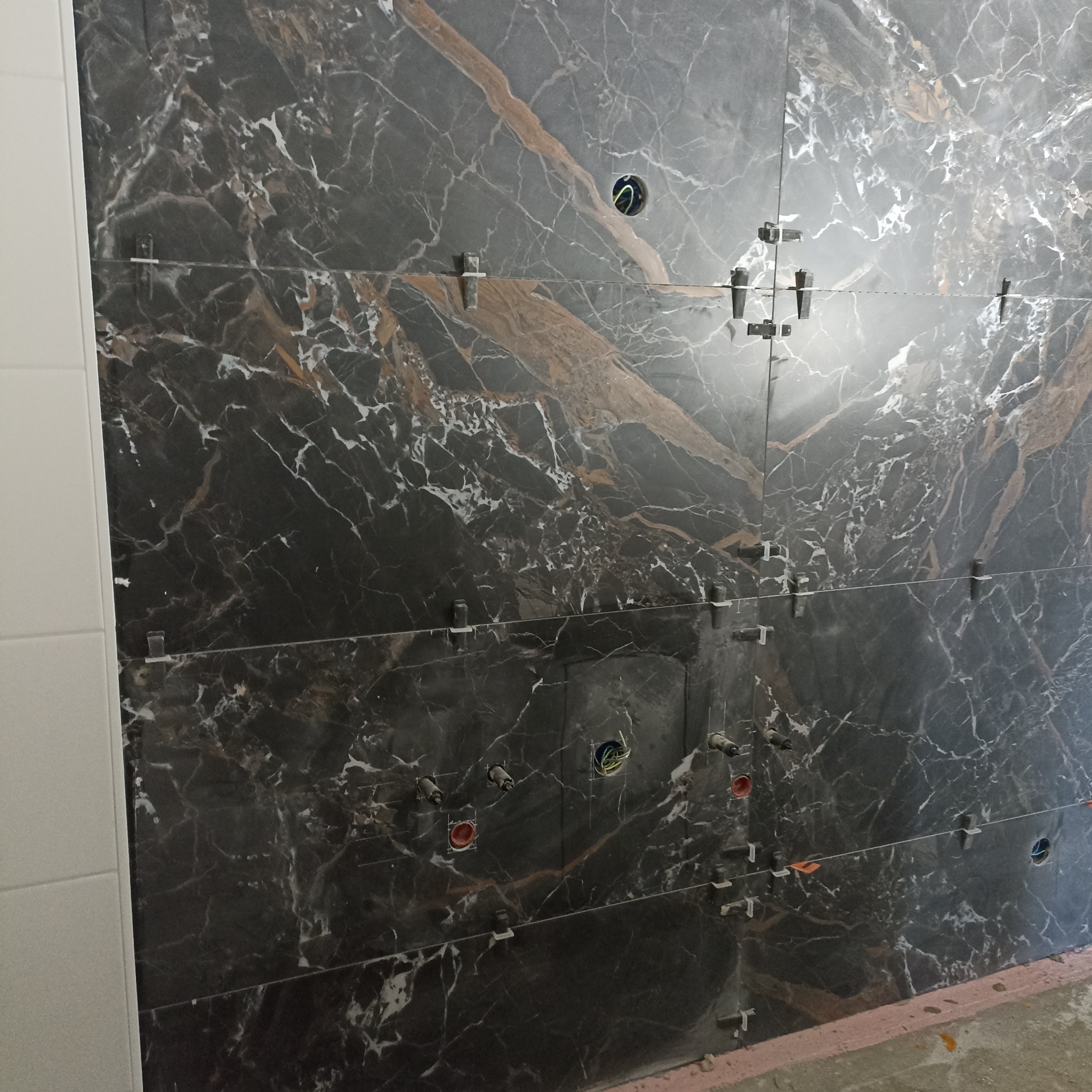 Układanie płytek imitujących marmur na ścianie z widocznymi otworami na instalacje elektryczne i sanitarną. Użyte krzyżyki dystansowe.