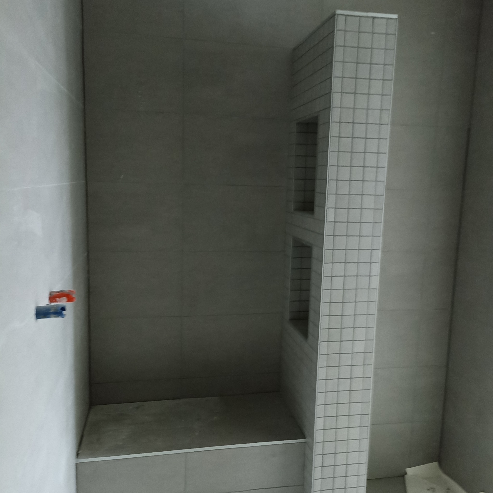 Wnęka prysznicowa w trakcie wykańczania. Widoczne płytki ceramiczne w odcieniach szarości, z małą wnęką na kosmetyki wyłożoną mozaiką. Instalacja wodna na ścianie.