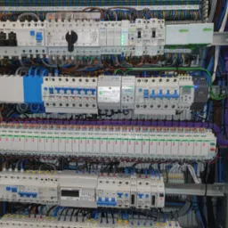 Rozbudowana instalacja elektryczna w szafie rozdzielczej, widoczne liczne bezpieczniki, przekaźniki i okablowanie. Elementy Schrack i Eaton.