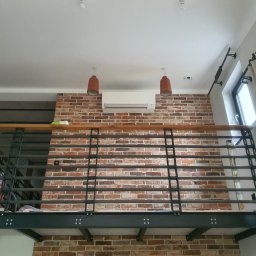 Recado - Loft z klimatyzacją na ceglanej ścianie, widok z dołu. Metalowa balustrada, wiszące lampy i okno z zasłoną. Czerwony odkurzacz na antresoli.