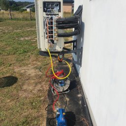 Recado - Montaż klimatyzacji zewnętrznej na białej ścianie budynku. Widoczne przewody, narzędzia i elementy instalacji. Trawa i drzewa w tle. Dzień słoneczny.