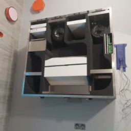 Centrala wentylacyjna z widocznymi filtrami i wentylatorami zamontowana na ścianie w pomieszczeniu z elementami instalacji elektrycznej.