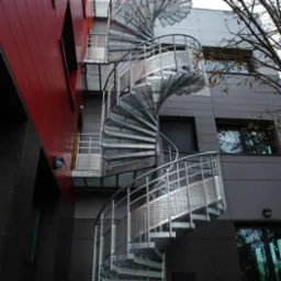 Zewnętrzne metalowe schody spiralne przy budynku o czerwono-szarej elewacji. Widok z dołu, ukazujący konstrukcję i detale schodów.