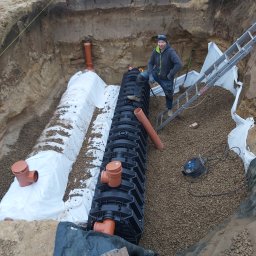 Recado - Instalacja kanalizacji deszczowej w wykopie ziemnym. Czarna, modułowa konstrukcja, rury PCV, biała geowłóknina. Mężczyzna stoi na drabinie w tle.