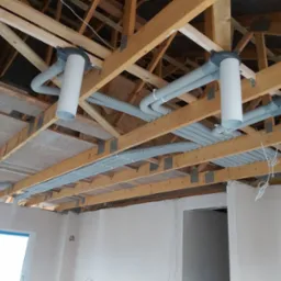 Wnętrze w trakcie montażu wentylacji mechanicznej: widoczne rury wentylacyjne pod stropem drewnianym, białe kanały wentylacyjne, surowy stan wykończenia ścian.