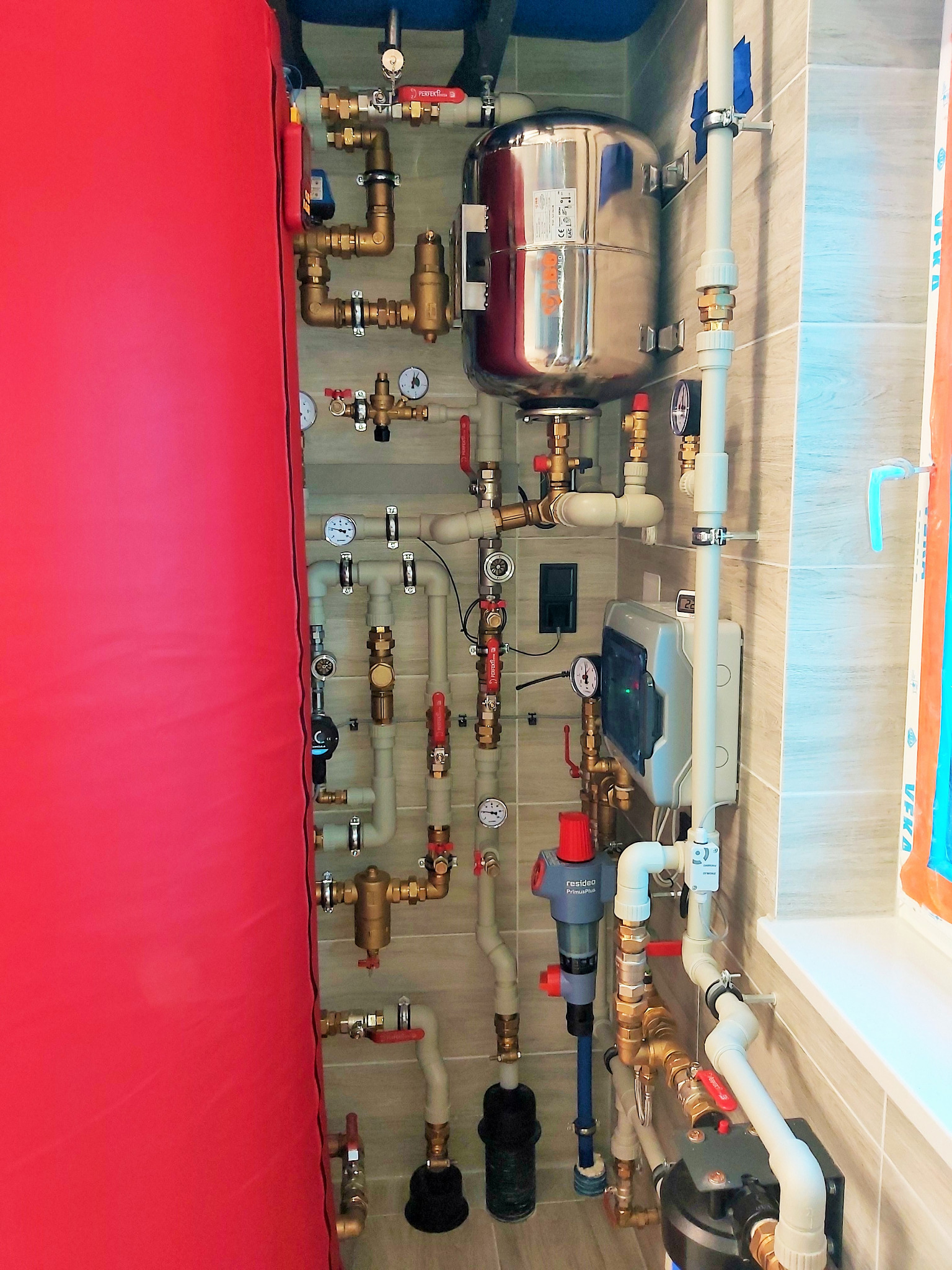 Kompleksowa instalacja hydrauliczna z licznymi rurami, zaworami i zbiornikiem wyrównawczym na tle wykończonej ściany z płytek. Widoczny filtr wody i elementy sterujące.